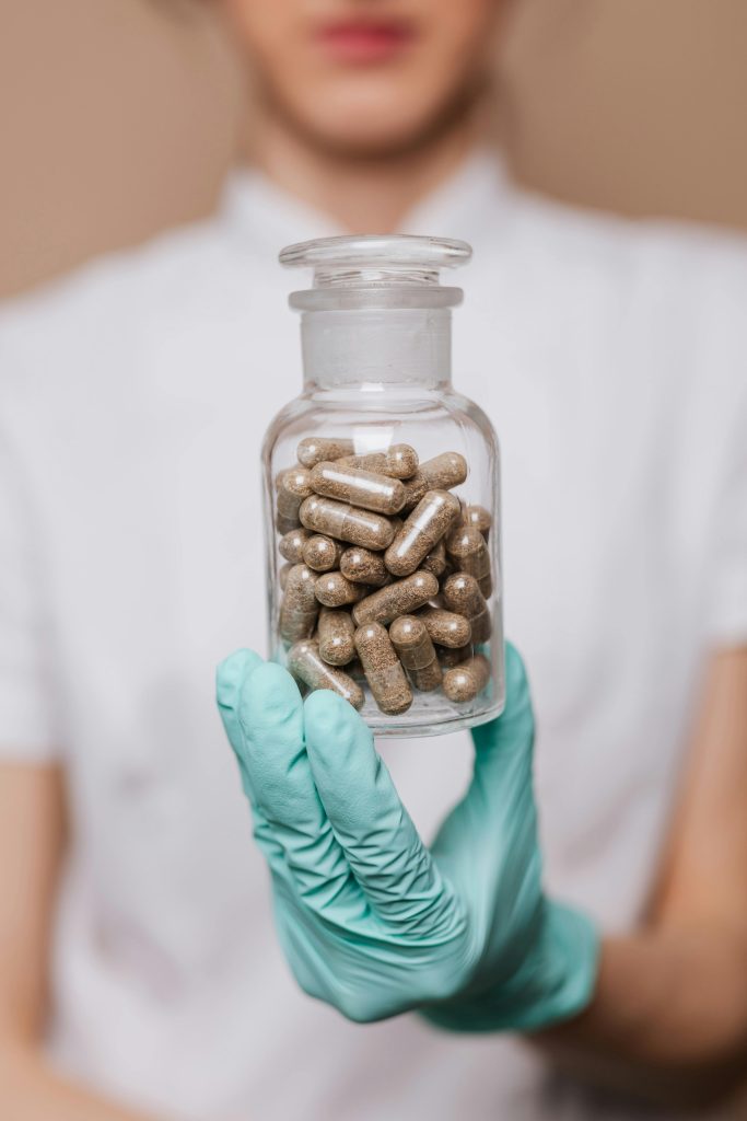 Ręka w turkusowej rękawiczce trzyma szklaną butelkę z brązowymi, ziołowymi kapsułkami. Koncept farmacji i suplementów.